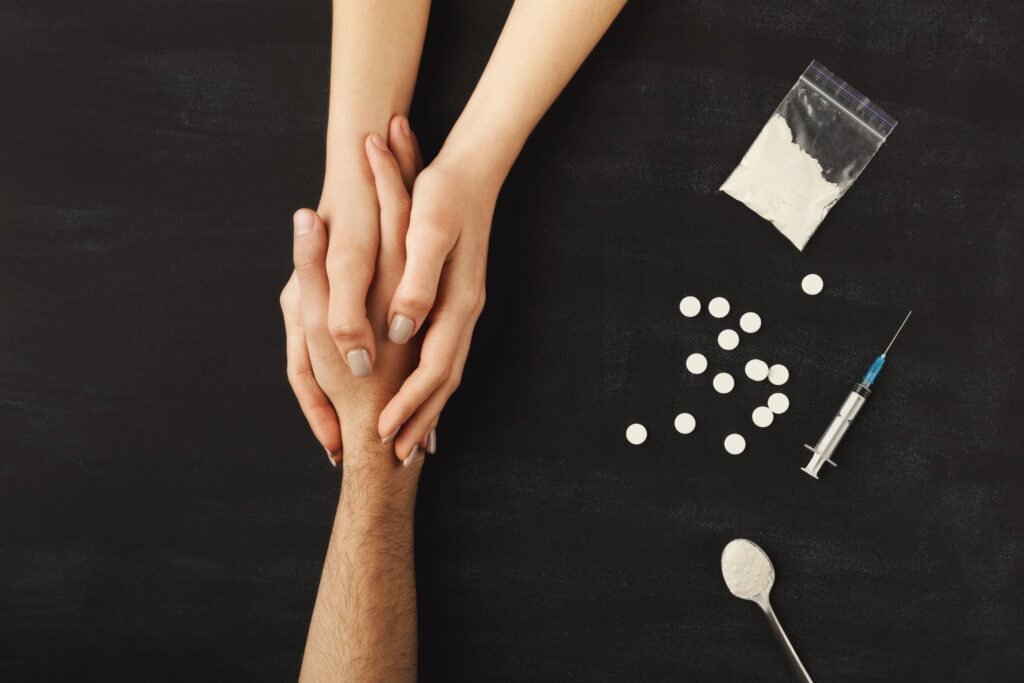 A person holding the hands of an individual struggling with fentanyl addiction, with pills and white powder on the table nearby, symbolizing support amidst the crisis.